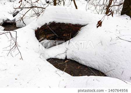 Winter's end stream flows around a snow-covered log. Thawed patches reveal dark water, hinting at spring's arrival amidst crisp white snow and bare branches 140764956