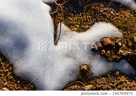 Sparkling snow patches melting on sun-warmed, rugged sandy-stony ground. A close-up of nature's crisp spring thaw, revealing earthy textures and tiny pebbles Sparkling snow patches melting on sun-warmed, rugged sandy-stony ground. A close-up of nature's crisp spring thaw, revealing earthy textures and tiny pebbles 140764975