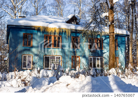 Charming weathered wooden house painted blue with heavy snow caps and long frozen icicles hanging from the roof during a bright and sunny winter day in the rural countryside 140765007