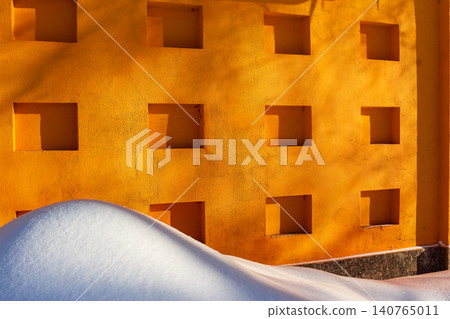 Large fluffy snowdrift rests against a vibrant orange wall featuring geometric recessed square niches under soft winter sunlight creating a striking minimalist contrast between cold and warmth Large fluffy snowdrift rests against a vibrant orange wall featuring geometric recessed square niches under soft winter sunlight creating a striking minimalist contrast between cold and warmth 140765011