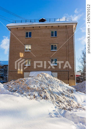 Old Soviet brick residential building stands behind a massive pile of snow in winter, clear blue sky background, typical Eastern European urban scene, cold sunny day lighting. Russia Old Soviet brick residential building stands behind a massive pile of snow in winter, clear blue sky background, typical Eastern European urban scene, cold sunny day lighting. Russia 140765012