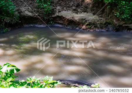 Turbid grey water flows through a muddy stream bed, visibly contaminated by industrial sewage discharge, creating an alarming scene of environmental pollution in a natural setting Turbid grey water flows through a muddy stream bed, visibly contaminated by industrial sewage discharge, creating an alarming scene of environmental pollution in a natural setting 140765032