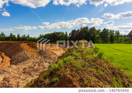 Open pit sand quarry scars the green field with massive mounds of soil and earth piles, showcasing raw industrial land modification against a vibrant forest backdrop under blue sky Open pit sand quarry scars the green field with massive mounds of soil and earth piles, showcasing raw industrial land modification against a vibrant forest backdrop under blue sky 140765040