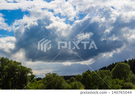 Massive dramatic storm clouds looming over vibrant green summer forest canopy, bright contrast between dark rain-bearing heavy cumulus and deep blue sky patches on a sunny afternoon Massive dramatic storm clouds looming over vibrant green summer forest canopy, bright contrast between dark rain-bearing heavy cumulus and deep blue sky patches on a sunny afternoon 140765044