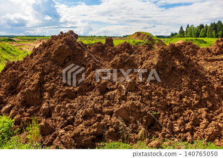 Large pile of raw dark soil mounds excavated from a field during construction work under a bright cloudy summer sky, showing natural clay and earth texture in an open landscape Large pile of raw dark soil mounds excavated from a field during construction work under a bright cloudy summer sky, showing natural clay and earth texture in an open landscape 140765050