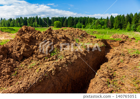 Deep trench dug into heavy clay soil on a construction site with large mounds of raw earth, contrasting with the lush green field and dense pine forest under a blue sky Deep trench dug into heavy clay soil on a construction site with large mounds of raw earth, contrasting with the lush green field and dense pine forest under a blue sky 140765051