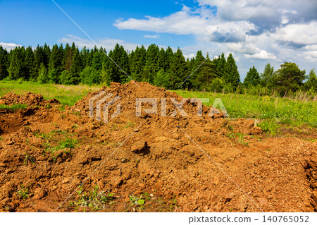 Large pile of raw earth dug up from a construction site in a green field, showing disturbed soil mounds and fresh dirt piles against a sunny landscape with pine trees in the distance Large pile of raw earth dug up from a construction site in a green field, showing disturbed soil mounds and fresh dirt piles against a sunny landscape with pine trees in the distance 140765052