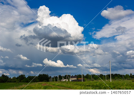 Dramatic fluffy cumulonimbus clouds floating across a vibrant deep blue summer sky over a scenic rural landscape with power lines stretching through a green meadow Dramatic fluffy cumulonimbus clouds floating across a vibrant deep blue summer sky over a scenic rural landscape with power lines stretching through a green meadow 140765057