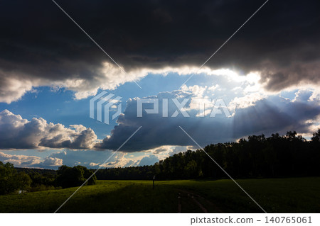 Dramatic sunbeams pierce through heavy storm clouds above a peaceful summer meadow, creating a powerful contrast between dark moody sky and bright divine light in the countryside 140765061