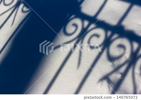 Elegant wrought iron fence shadow forms a striking abstract pattern on fresh white snow. A bright winter day creates this beautiful, graphic nature background Elegant wrought iron fence shadow forms a striking abstract pattern on fresh white snow. A bright winter day creates this beautiful, graphic nature background 140765072