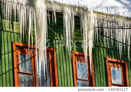 Sunlit, sharp icicles pierce the winter sky, hanging from an aged green timber dwelling. Bright contrast against the weathered facade, Eastern Europe Sunlit, sharp icicles pierce the winter sky, hanging from an aged green timber dwelling. Bright contrast against the weathered facade, Eastern Europe 140765127