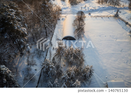 Aerial drone shot: a snow-covered arch hangar anchors a vast, fenced winter field. Golden sunlight warms the pristine, frosty landscape, creating striking shadows and a tranquil scene Aerial drone shot: a snow-covered arch hangar anchors a vast, fenced winter field. Golden sunlight warms the pristine, frosty landscape, creating striking shadows and a tranquil scene 140765130
