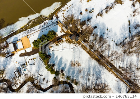 Stunning aerial view: a serene snowy riverbank retreat with a large wooden deck. Melting ice drifts on the murky river. Long tree shadows pattern the pristine white winter landscape Stunning aerial view: a serene snowy riverbank retreat with a large wooden deck. Melting ice drifts on the murky river. Long tree shadows pattern the pristine white winter landscape 140765134