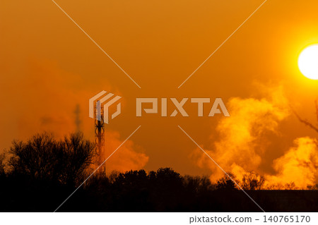 Towering industrial stack emits sunlit steam and smoke into a vivid orange sky during a breathtaking winter sunset Towering industrial stack emits sunlit steam and smoke into a vivid orange sky during a breathtaking winter sunset 140765170