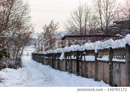 Snow-covered concrete fence with menacing razor wire coil, delineating a bleak industrial zone boundary in winter. A snowy path leads into the distance Snow-covered concrete fence with menacing razor wire coil, delineating a bleak industrial zone boundary in winter. A snowy path leads into the distance 140765172
