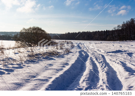 Winding tire tracks carve through a pristine snow-covered field on a bright winter's day. A striking lone tree and distant forest under a serene blue sky Winding tire tracks carve through a pristine snow-covered field on a bright winter's day. A striking lone tree and distant forest under a serene blue sky 140765183