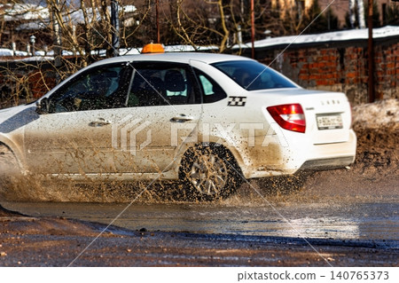 White taxi car splashes through a muddy spring puddle, sending up a vibrant spray of dirty water and slush on a thawing road 140765373