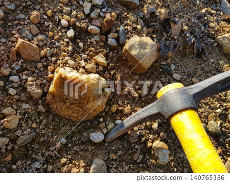 Rock hammer and intriguing raw mineral on pebbly earth. Geologist's find, showcasing earth science and exploration discovery Rock hammer and intriguing raw mineral on pebbly earth. Geologist's find, showcasing earth science and exploration discovery 140765386