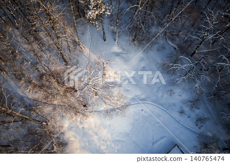 Aerial winter panorama: a lone figure's silhouette trails a winding path through a snow-covered forest. Golden sunlight illuminates frosty branches and deep shadows Aerial winter panorama: a lone figure's silhouette trails a winding path through a snow-covered forest. Golden sunlight illuminates frosty branches and deep shadows 140765474