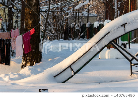 Deserted children's slide blanketed by fresh, pristine snow, glowing under soft winter sunshine. A silent, frosty scene, evoking peaceful cold 140765490