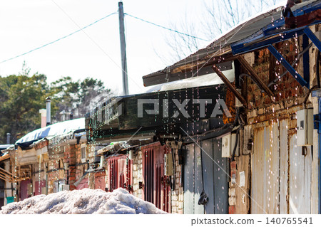 Melting snow dripping from rusty garage rooftops during a sunny spring thaw, capturing the seasonal transition and water droplets falling from metal structures under the bright sky Melting snow dripping from rusty garage rooftops during a sunny spring thaw, capturing the seasonal transition and water droplets falling from metal structures under the bright sky 140765541