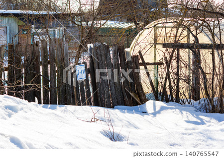 Rustic wooden picket fence with a numbered gate in a quiet countryside garden setting during the thaw of early spring with patches of melting snow and an arched greenhouse nearby Rustic wooden picket fence with a numbered gate in a quiet countryside garden setting during the thaw of early spring with patches of melting snow and an arched greenhouse nearby 140765547