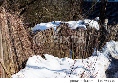 Dilapidated weathered wooden fence covered with dry climbing vines in a rustic garden during early spring thaw with melting snow patches and rural house background Dilapidated weathered wooden fence covered with dry climbing vines in a rustic garden during early spring thaw with melting snow patches and rural house background 140765550