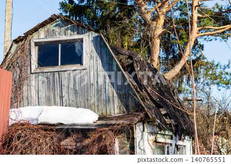 Rustic old wooden house with a weathered facade and damaged roof under spring sun, surrounded by bare trees and melting snow patches in a neglected suburban cottage garden 140765551