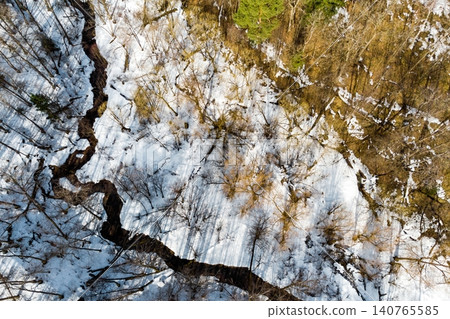 Winding stream through a thawing, snow-dusted forest, seen from above. Bare trees cast long shadows on melting patches, signaling early spring's arrival Winding stream through a thawing, snow-dusted forest, seen from above. Bare trees cast long shadows on melting patches, signaling early spring's arrival 140765585