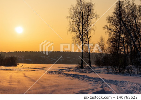 Golden winter field bathed in warm evening sun. Snow-covered path leads towards bare trees, silhouetted against an orange sky. Peaceful frosty landscape Golden winter field bathed in warm evening sun. Snow-covered path leads towards bare trees, silhouetted against an orange sky. Peaceful frosty landscape 140765622