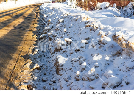 High snow banks line a cleared winter road surface glistening under bright sun, showing signs of plowing activity High snow banks line a cleared winter road surface glistening under bright sun, showing signs of plowing activity 140765665