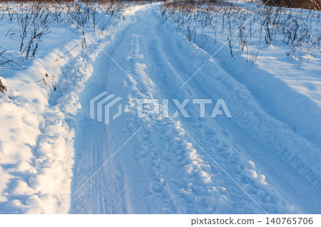 Winding snowy path etched with tire tracks and footprints, bathed in winter light. Pristine white snow and frozen vegetation evoke serene natural beauty Winding snowy path etched with tire tracks and footprints, bathed in winter light. Pristine white snow and frozen vegetation evoke serene natural beauty 140765706