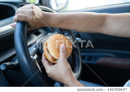 Asian lady holding hamburger to eat in car, dangerous and risk an accident. Asian lady holding hamburger to eat in car, dangerous and risk an accident. 140769647
