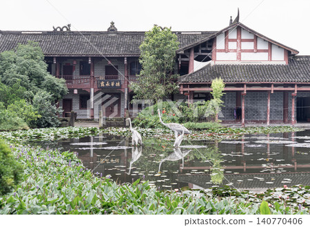 Before the weathered tile roofs and traditional wooden balconies of a grand Chinese estate, a glassy pond rests in perfect stillness. Flanked by lush foliage and floating lotus pads, a group of Before the weathered tile roofs and traditional wooden balconies of a grand Chinese estate, a glassy pond rests in perfect stillness. Flanked by lush foliage and floating lotus pads, a group of 140770406