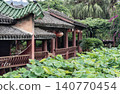 An elegant wooden pavilion with sweeping green-tiled roofs curves gracefully beside a vibrant pond filled with lush lotus leaves. Strung beneath the low eaves, crimson lanterns add a warm touch 140770454