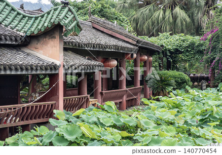 An elegant wooden pavilion with sweeping green-tiled roofs curves gracefully beside a vibrant pond filled with lush lotus leaves. Strung beneath the low eaves, crimson lanterns add a warm touch An elegant wooden pavilion with sweeping green-tiled roofs curves gracefully beside a vibrant pond filled with lush lotus leaves. Strung beneath the low eaves, crimson lanterns add a warm touch 140770454