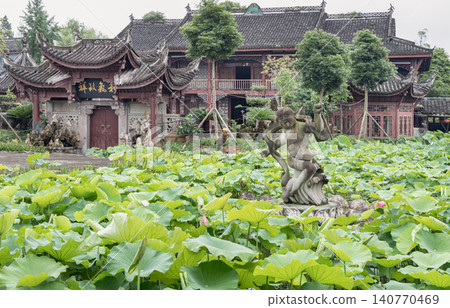 A weathered stone statue rises gracefully from a sprawling sea of vibrant green lotus leaves, capturing a dynamic, fluid motion amidst the stillness of the pond. In the background, the intricate, 140770469