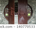 Framed by a weathered grey stone wall, a traditional Chinese moon gate stands partially open, its heavy crimson wooden doors ajar. The central opening reveals a recessed stone corridor leading to a 140770533