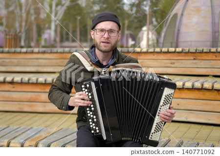 An alcoholic accordion player plays the accordion in the park. A man with glasses is sitting on a bench, holding an accordion in his hands. The soulful musician sings songs An alcoholic accordion An alcoholic accordion player plays the accordion in the park. A man with glasses is sitting on a bench, holding an accordion in his hands. The soulful musician sings songs An alcoholic accordion 140771092