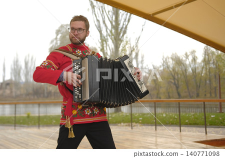 Portrait of an authentic musician in national costume playing the accordion in the park. A white man with glasses is playing a new tune on the accordion. An outdoor scene with a river Portrait of an authentic musician in national costume playing the accordion in the park. A white man with glasses is playing a new tune on the accordion. An outdoor scene with a river 140771098