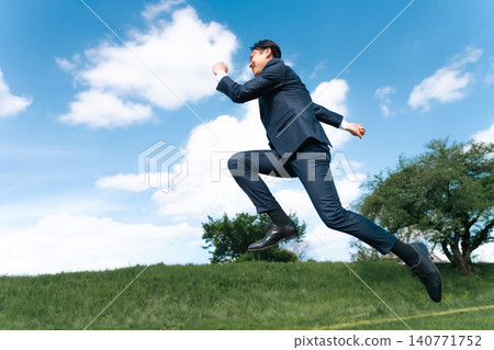 A Japanese businessman in a suit walks briskly across a grassland under a blue sky. A Japanese businessman in a suit walks briskly across a grassland under a blue sky. 140771752