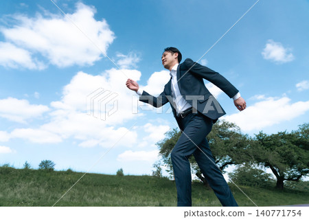 A Japanese businessman in a suit walks briskly across a grassland under a blue sky. A Japanese businessman in a suit walks briskly across a grassland under a blue sky. 140771754
