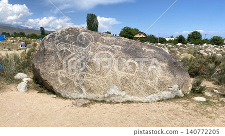 The Museum of petroglyphs on Issyk-Kul in Kyrgyzstan. View of a large boulder with prehistoric drawings of animals and hunting scenes. snow-capped mountain peaks in the distance The Museum of petroglyphs on Issyk-Kul in Kyrgyzstan. View of a large boulder with prehistoric drawings of animals and hunting scenes. snow-capped mountain peaks in the distance 140772205