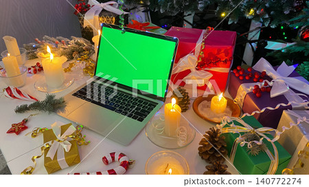 Festive New Year's table with a laptop with a chroma key on the screen. Colorful gift boxes, burning candles, cones, confetti and other elements of Christmas. Festive New Year's table with a laptop with a chroma key on the screen. Colorful gift boxes, burning candles, cones, confetti and other elements of Christmas. 140772274