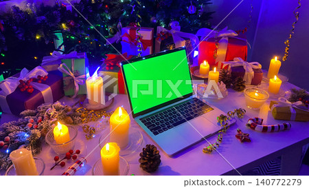 New Year computer concept. Laptop with a chroma key on the screen lies on the Christmas table. Wrapped gift boxes, burning candles, pine cones, confetti, fir branch with red berries 140772279
