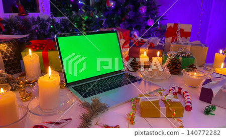 Laptop with a chroma key on the screen is on the New Year's table. Beautifully packaged gift boxes, burning candles, cones, confetti, a fir branch with red berries and striped caramel Laptop with a chroma key on the screen is on the New Year's table. Beautifully packaged gift boxes, burning candles, cones, confetti, a fir branch with red berries and striped caramel 140772282