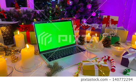 Personal computer with a chroma key on the screen lies on the New Year's table. Freelancer's Christmas workplace. Gift boxes, burning candles, cones, confetti, a fir branch with red berries Personal computer with a chroma key on the screen lies on the New Year's table. Freelancer's Christmas workplace. Gift boxes, burning candles, cones, confetti, a fir branch with red berries 140772294