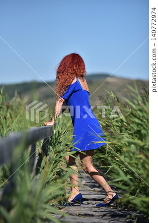 Woman with red hair in blue dress standing on wooden footbridge in a reed field Woman with red hair in blue dress standing on wooden footbridge in a reed field 140772974
