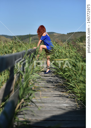 Woman with red hair showcases a vivid blue dress while standing on a wooden footbridge amidst reed Woman with red hair showcases a vivid blue dress while standing on a wooden footbridge amidst reed 140772975