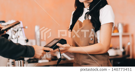 Asian senior female barista adult receiving mobile payment from customer using smartphone at cafe demonstrating modern cashless transaction in cozy local family coffee shop Asian senior female barista adult receiving mobile payment from customer using smartphone at cafe demonstrating modern cashless transaction in cozy local family coffee shop 140773451